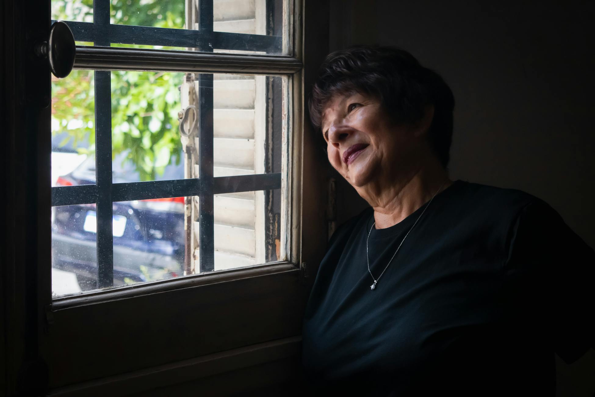 A senior woman looking thoughtfully out a window, framed by soft light and shadows.