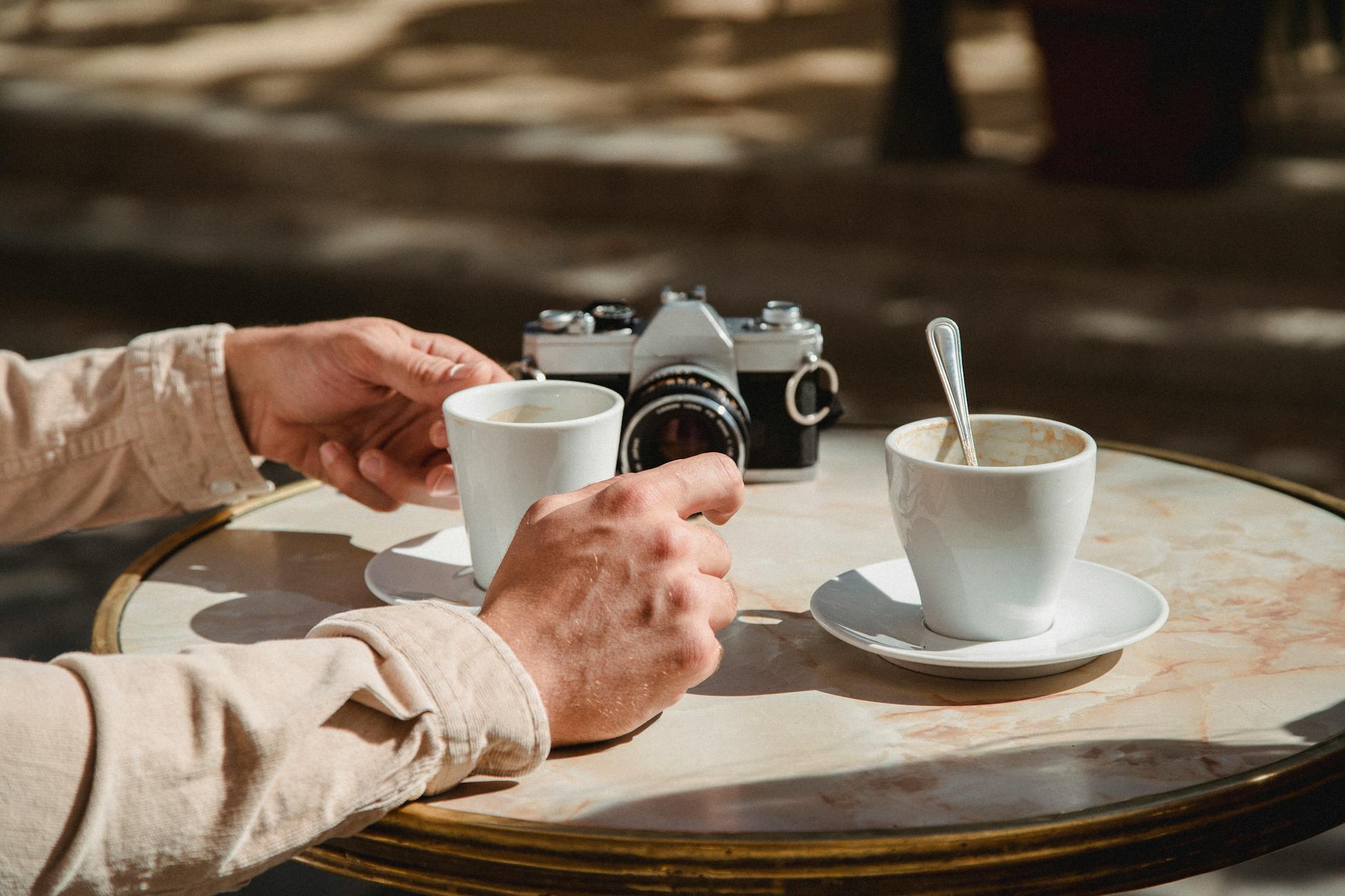 Hands holding coffee cups outdoors with a vintage camera on a table.