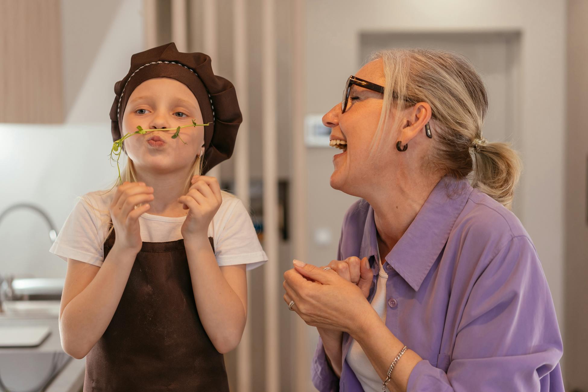 A senior woman and her young grandchild enjoy playful cooking fun indoors, sharing smiles and laughter.