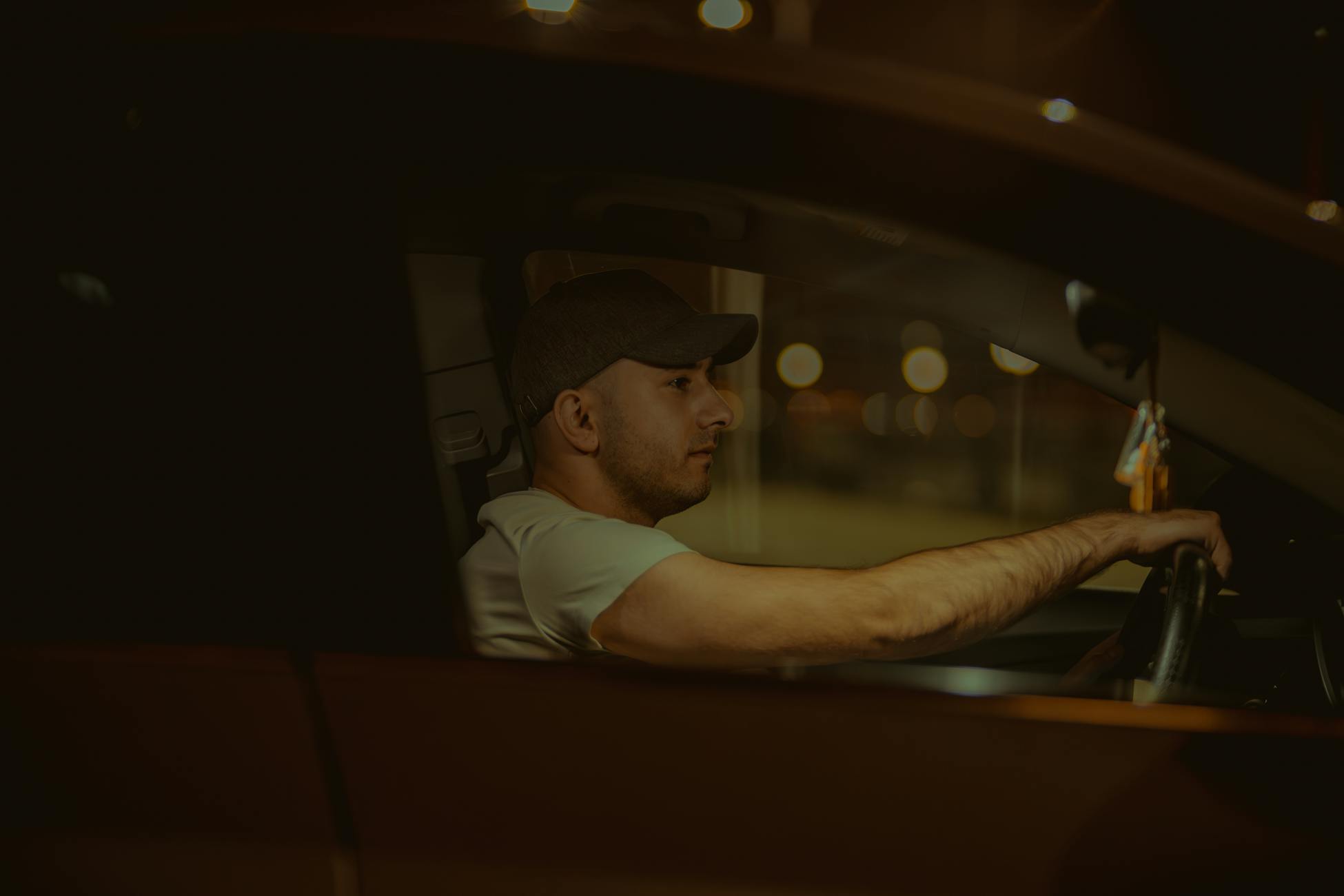 A man wearing a cap is driving a car at night, focused on the road ahead.