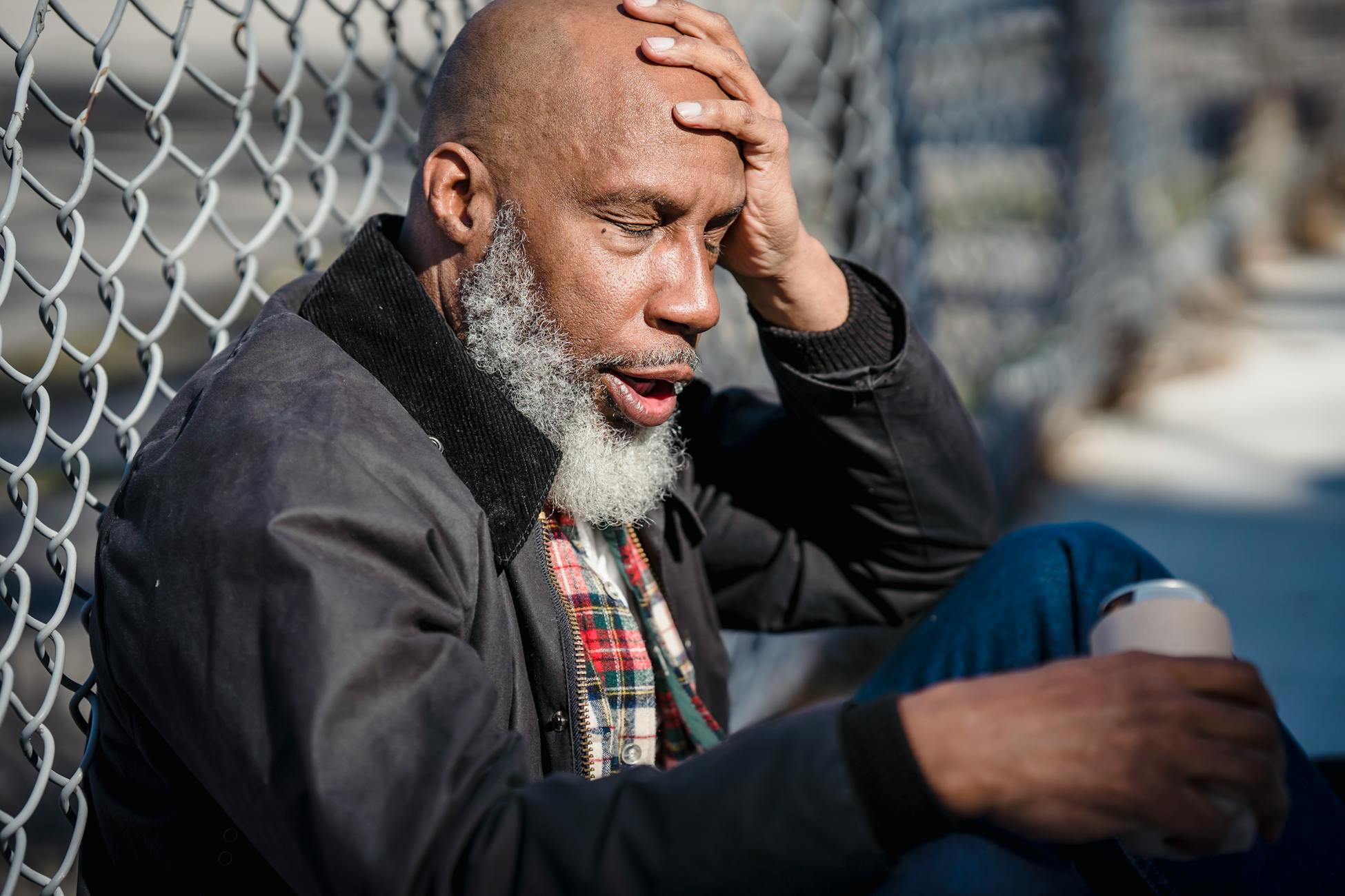 An older man with a beard holding a drink, seated by a fence outdoors.