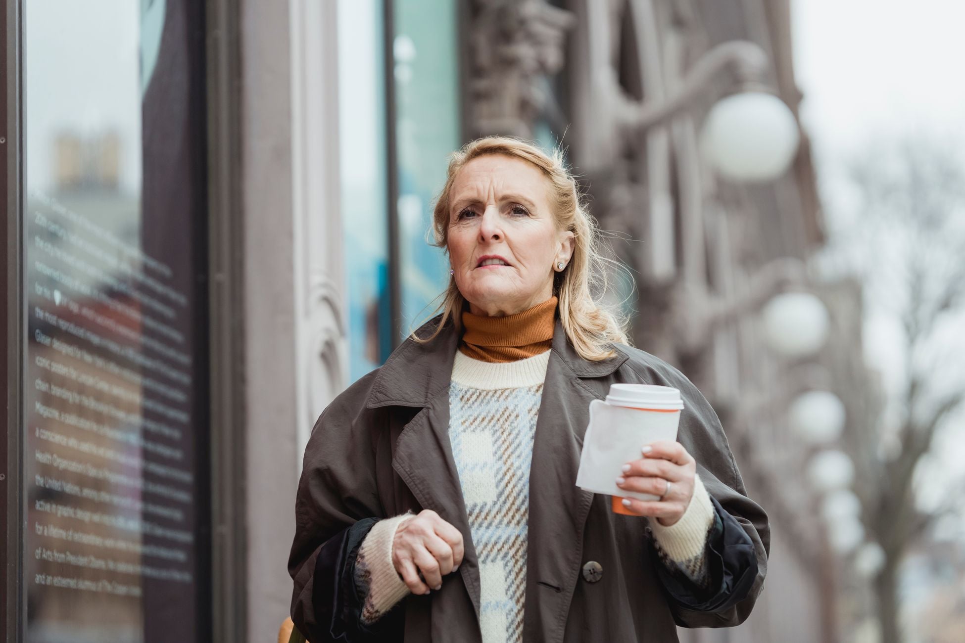 Elderly female in coat with disposable cup of hot beverage looking forward in city in daytime
