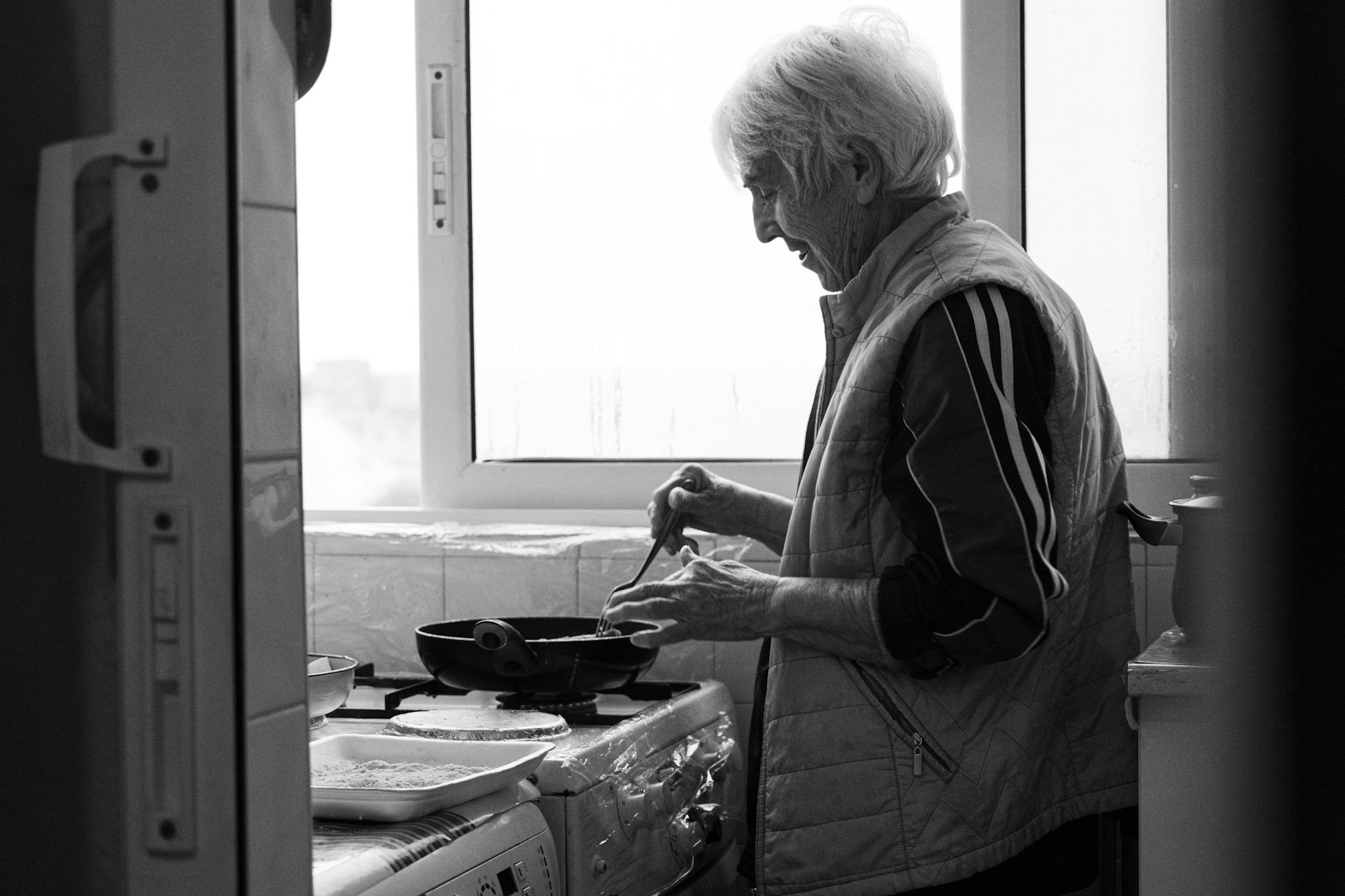 Elderly woman cooking at home, capturing warmth and tradition in a black and white setting.