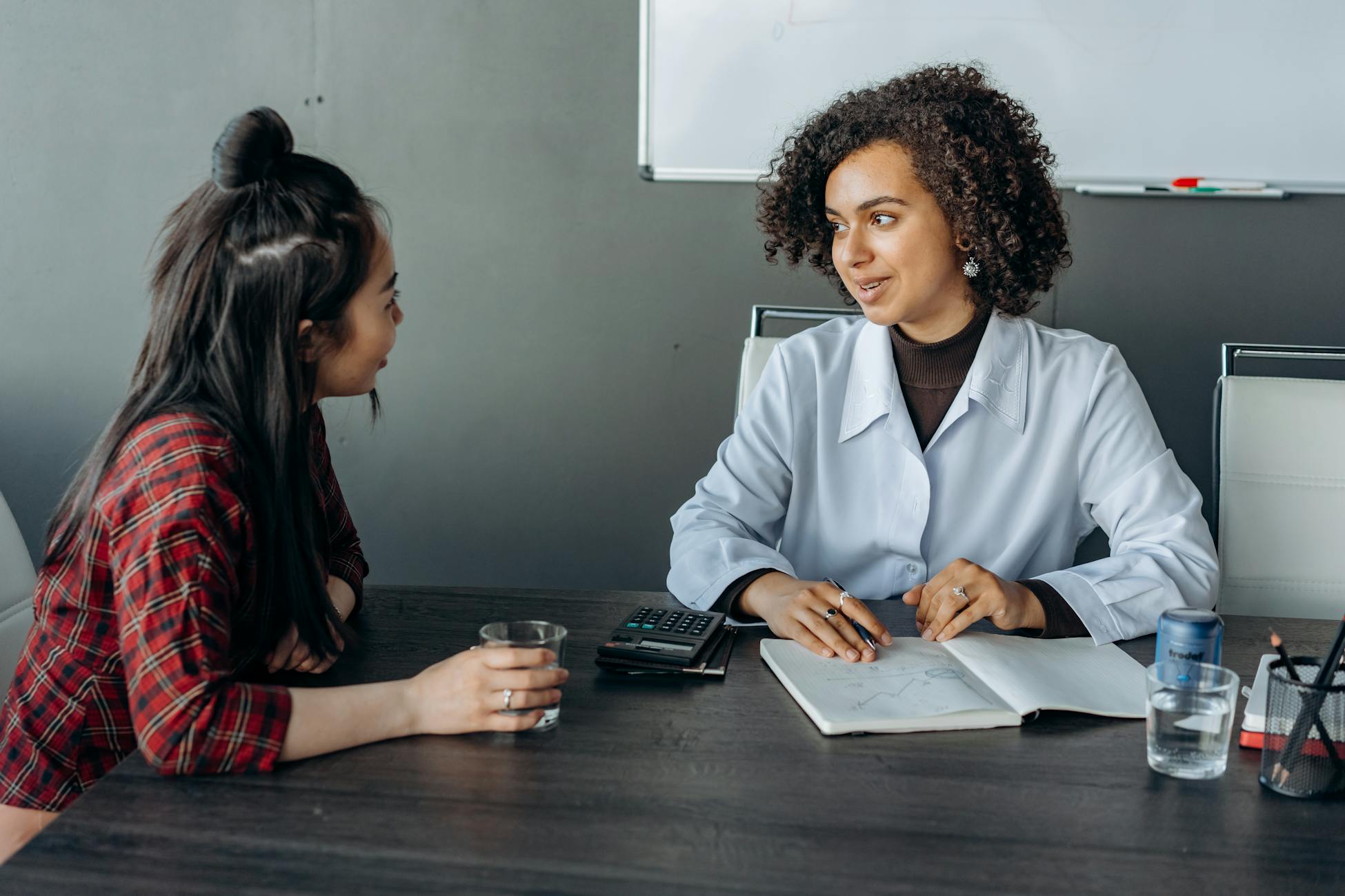 Two women in an office setting discussing work across a wooden table.