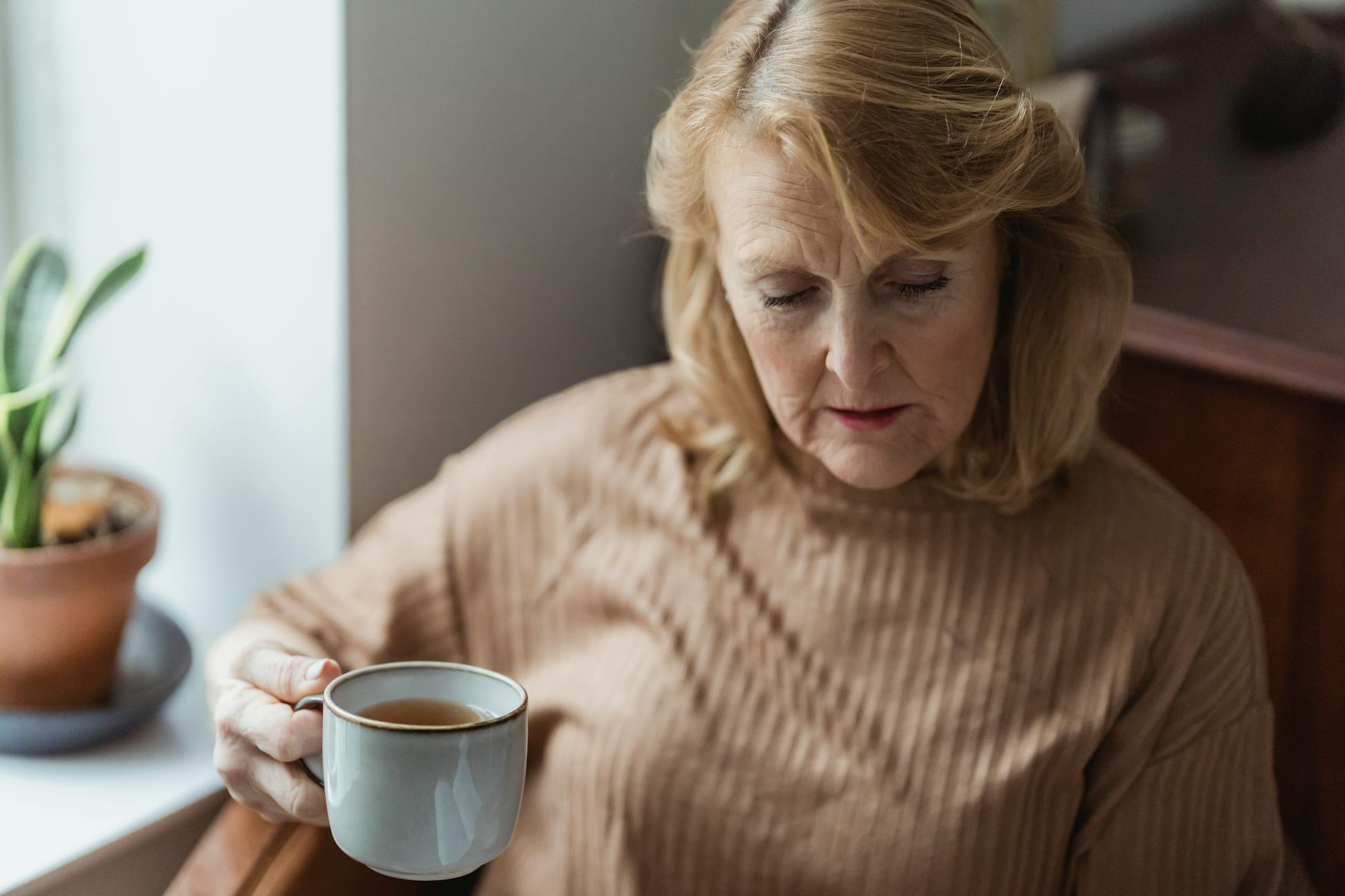 Crop focused elderly lady drinking cup of hot tea and looking down while resting near window at home